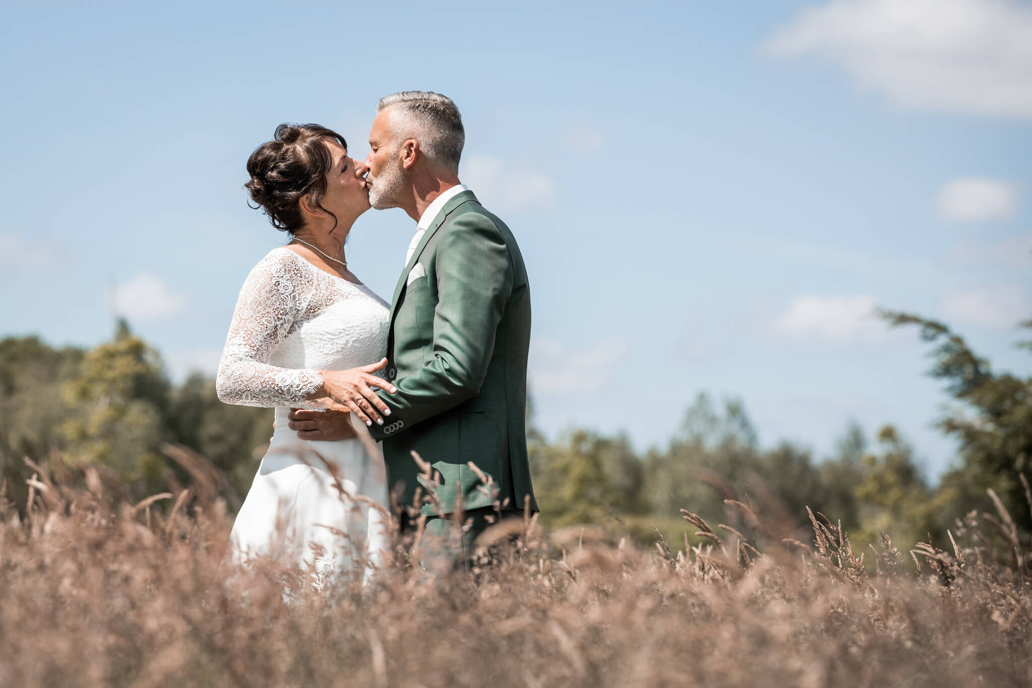 Bruidspaar kust elkaar in hoog grasland in mooie groene omgeving tijdens de fotoshoot op hun trouwdag.